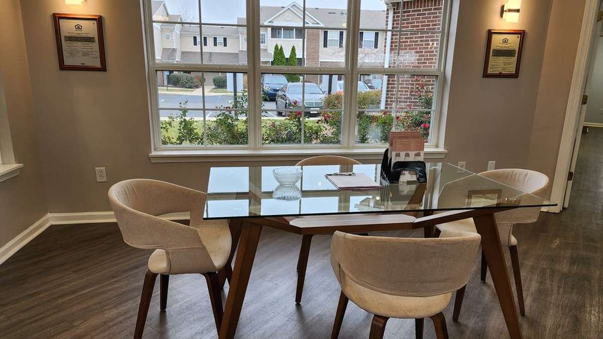 A bright, clean clubhouse dining room at an apartment community — a glass table and upholstered chairs by a large window looking onto the buildings outside.